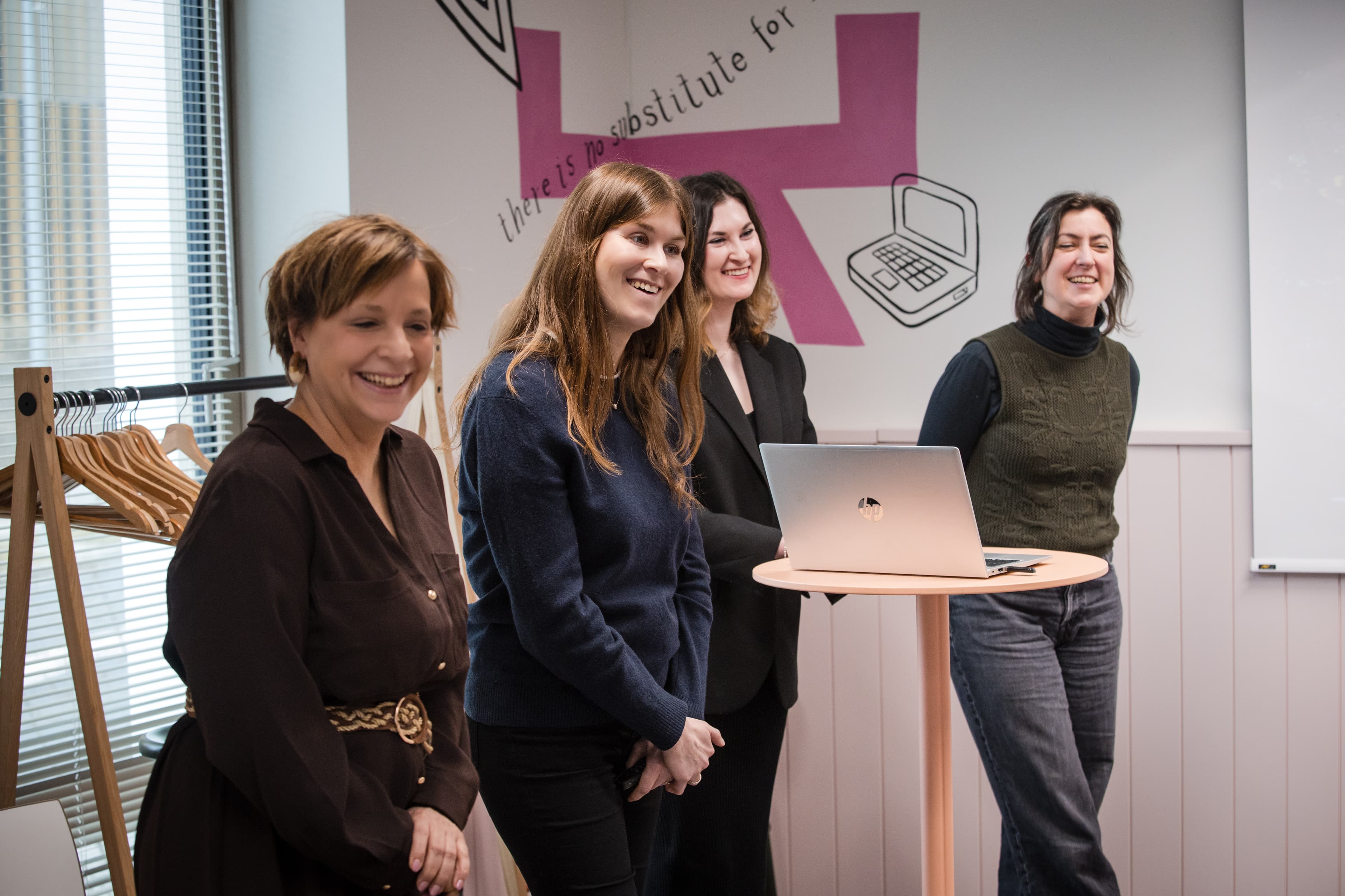 Four women smiling around a laptop on a high table in a colourful creative room