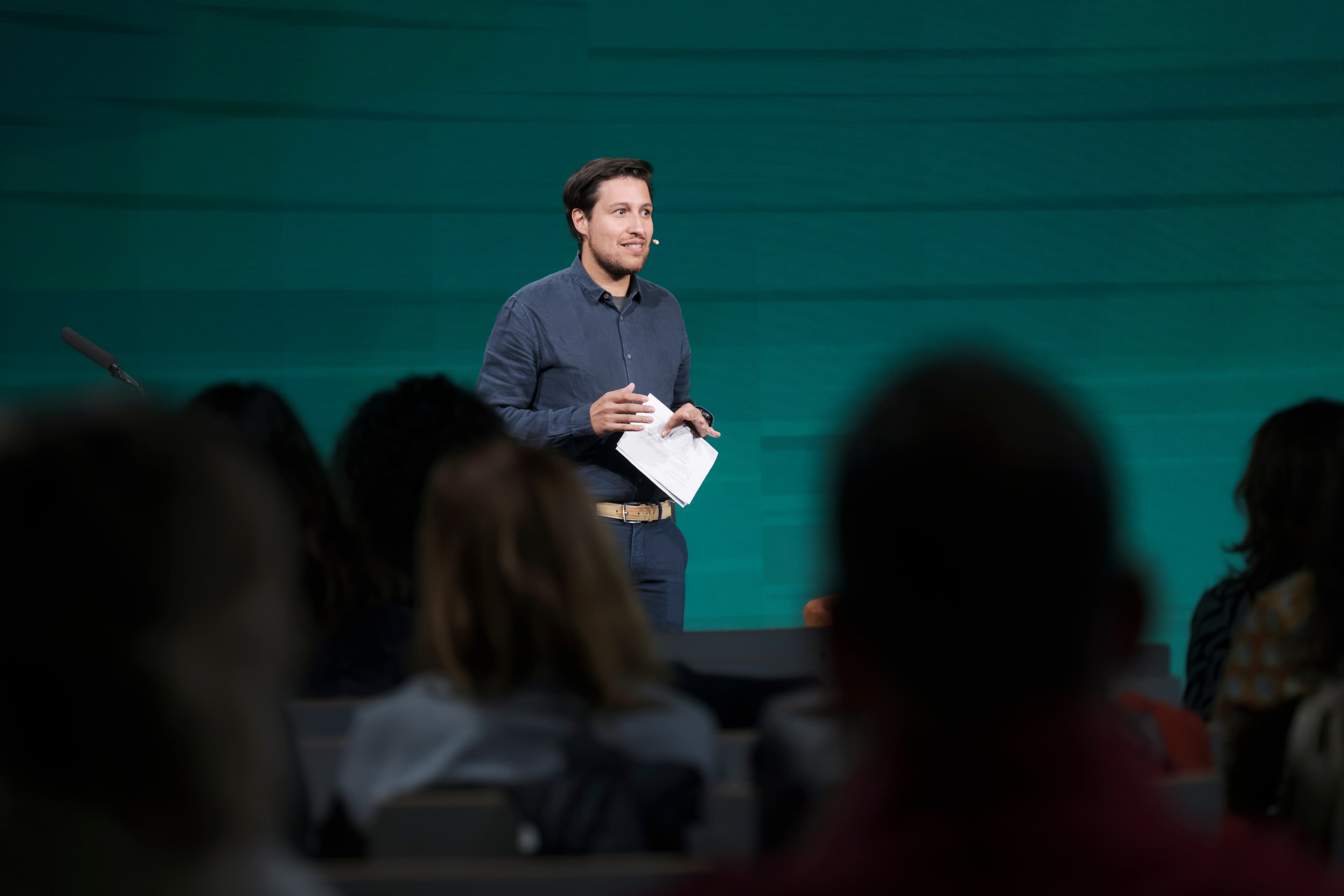 A speaker holding notes addresses the audience on stage against a green backdrop