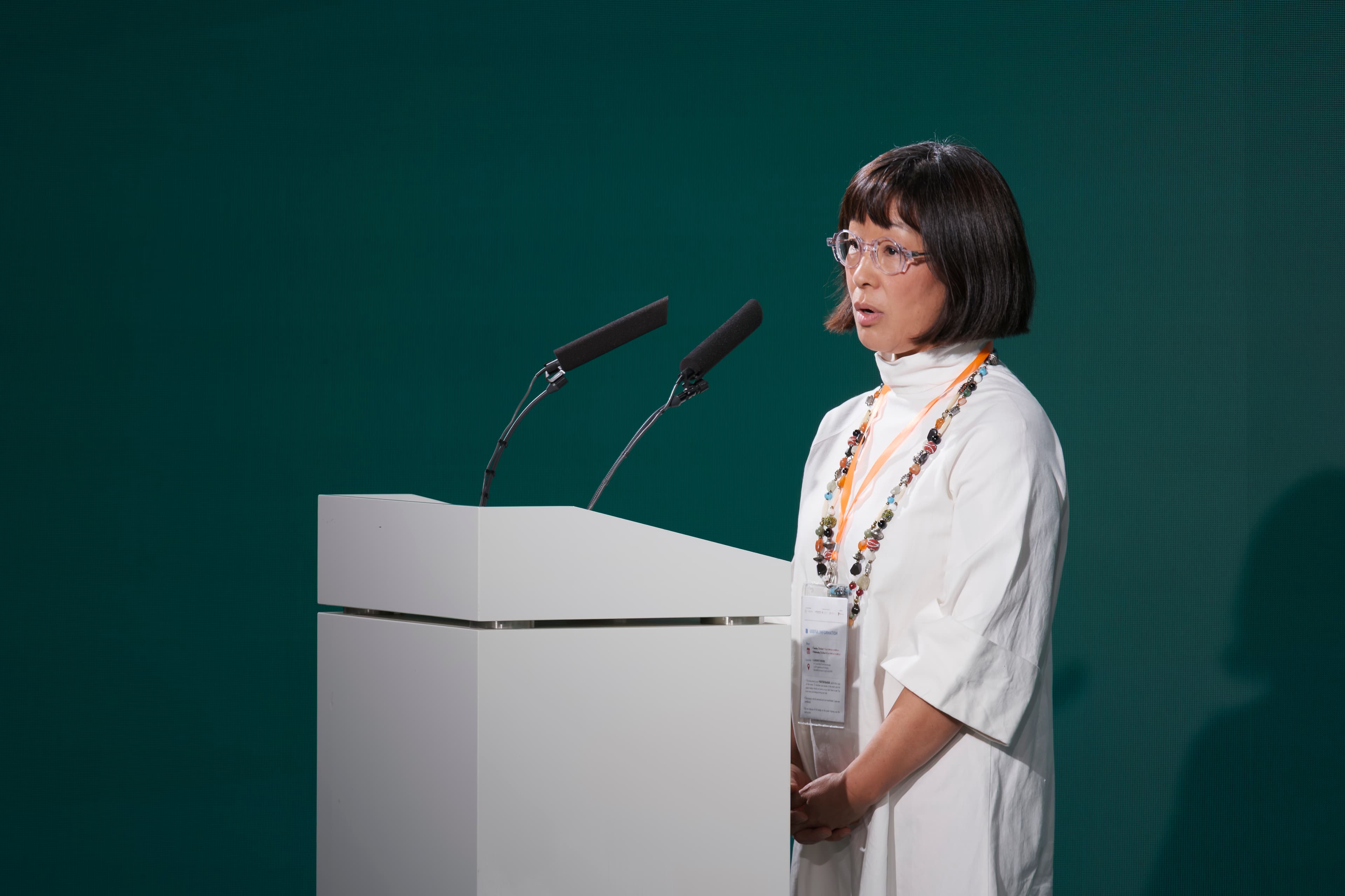A woman in white speaks at a lectern against a green backdrop at the SPX Colloquium
